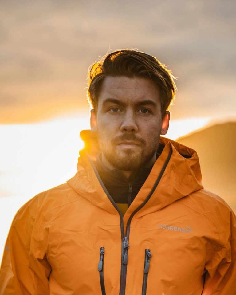 A man in a bright jacket posing outdoors at sunset, showcasing modern adventure style.