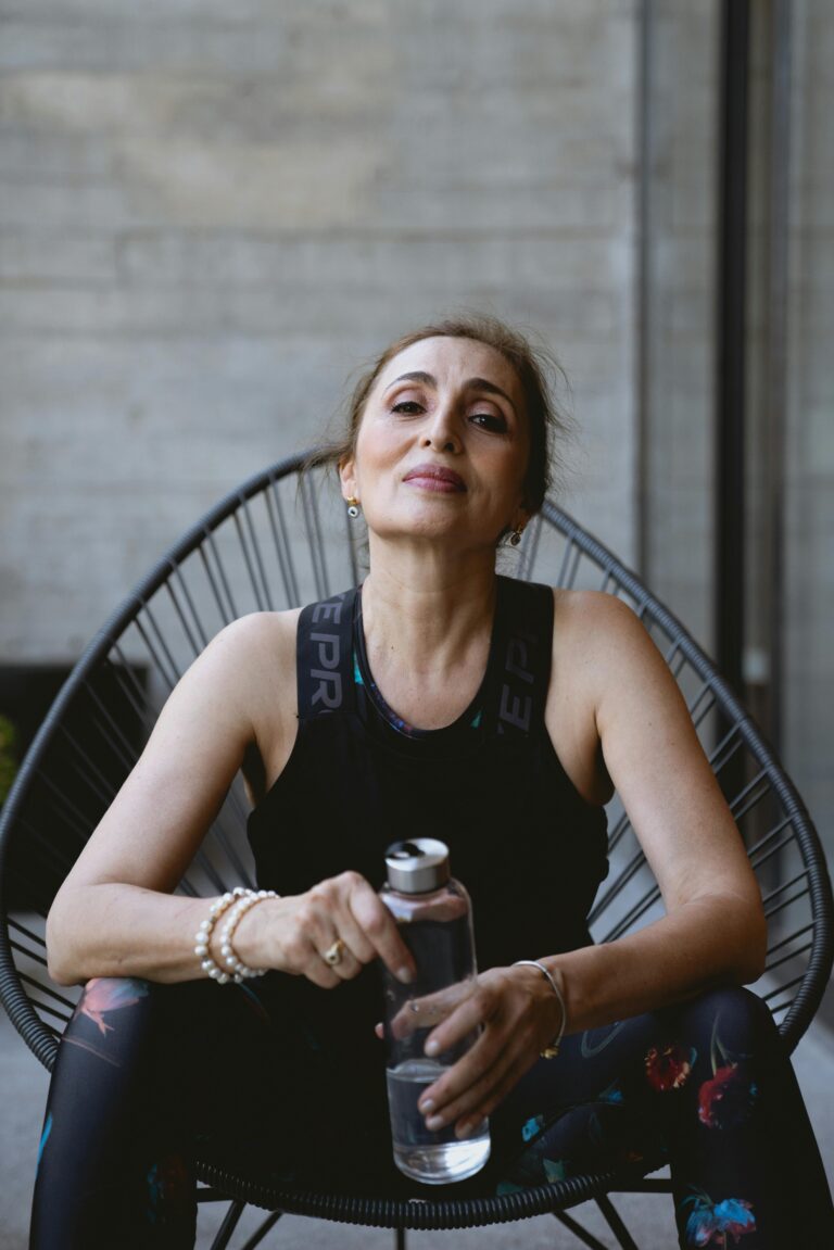 Mature woman in a black tank top holding a water bottle and resting indoors after exercising.